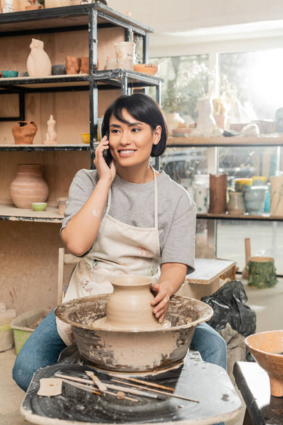 Vrolijke jonge Aziatische ambachtsman in schort praten op smartphone en zitten in de buurt van klei op aardewerk wiel en houten gereedschappen in keramische werkplaats, aardewerk creatie proces - Foto, afbeelding