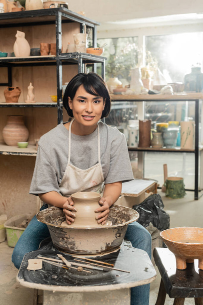 Lächelnde junge asiatische Kunsthandwerkerin in Schürze, die die Form einer Tonvase herstellt und in die Kamera schaut, während sie mit der Töpferscheibe in einem verschwommenen Kunstworkshop arbeitet, Töpferei-Entstehungsprozess - Foto, Bild