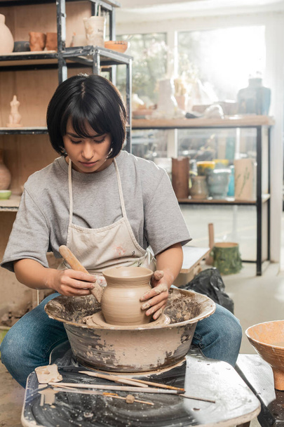 Jonge brunette aziatische vrouwelijke pottenbakker in schort met houten gereedschap in de buurt van klei, terwijl het werken met draaiende aardewerk wiel in wazig keramiek workshop, aardewerk creatie proces - Foto, afbeelding
