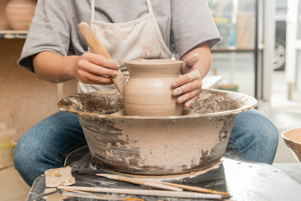 エプロンの若い女性職人の作物のビューは、バックグラウンドでぼやけたセラミックワークショップで陶芸ホイールを回転させる上で木製のツールと粘土花瓶の形を作る、陶器の作成プロセス - 写真・画像