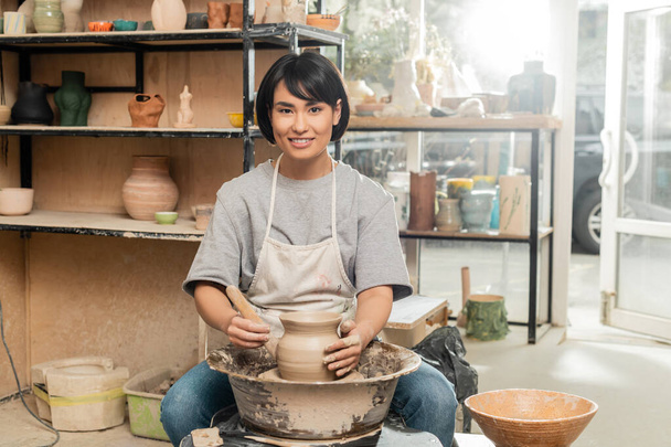 Sonriente joven morena asiática artesana en delantal mirando a la cámara mientras sostiene la herramienta de madera cerca de la arcilla en la rueda de cerámica en el estudio de cerámica en el fondo, moldeado de arcilla y proceso de formación - Foto, imagen