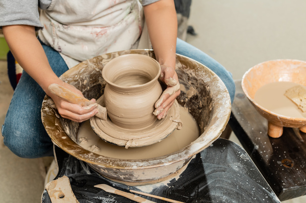 Widok upraw młodej garncarki w fartuchu kształtowania wazonu glinianego z drewnianym narzędziem na kole garncarskim w pobliżu miski z wodą i gąbką w pracowni sztuki, kształtowania gliny i procesu formowania - Zdjęcie, obraz