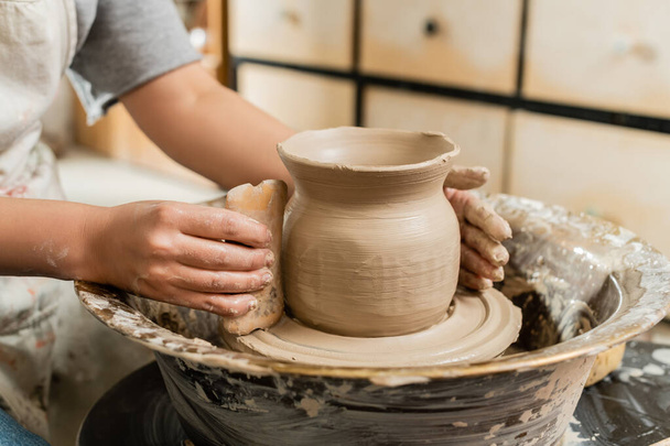 陶片に木製のスクレーパーで粘土の花瓶を作るエプロンの若い女性職人の作物のビューは、ぼやけたセラミックワークショップ、粘土の形成と形成プロセス - 写真・画像
