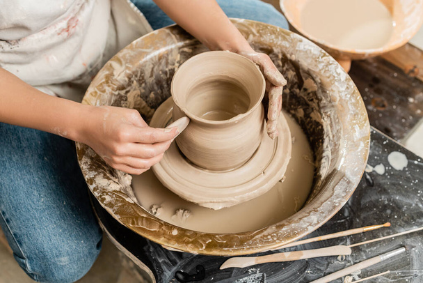 Top view of young female potter in apron making shape of clay vase with wooden tool on spinning pottery tool in ceramic workshop, clay shaping and forming process - Foto, Imagem