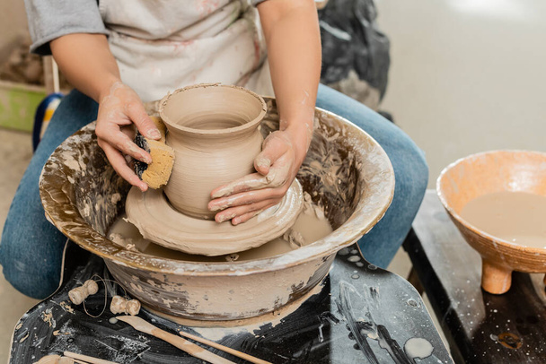Widok upraw garncarki w fartuchu kształtowania wazonu glinianego z gąbką weterynaryjną w pobliżu drewnianych narzędzi i kręcące się koło ceramiczne w warsztacie ceramicznym, kształtowanie gliny i proces formowania - Zdjęcie, obraz
