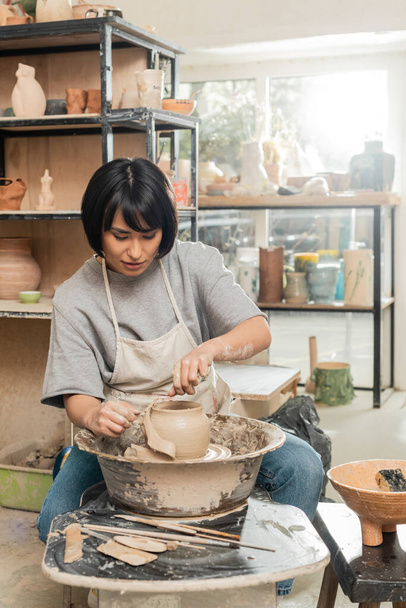 Brunetka azjatyckie kobieta rzemieślnik w fartuch cięcia mokrej gliny na przędzenia ceramiki koła podczas pracy w pobliżu drewnianych narzędzi i miski w niewyraźne warsztat ceramiczny, kształtowanie gliny i procesu formowania - Zdjęcie, obraz