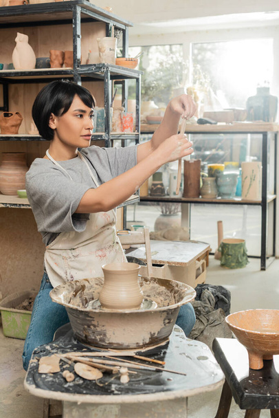 Молода брюнетка азіатська жінка-гончарка в фартусі тримає глину біля вази на обертанні гончарного колеса біля дерев'яних інструментів і чаші в розмитій керамічній майстерні, виробництво і процес промислового гончарства - Фото, зображення