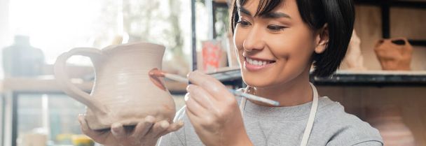 Smiling young asian female artisan painting on clay jug while working in blurred ceramic workshop at background, clay shaping technique and process, banner  - 写真・画像