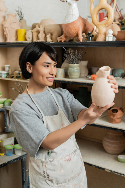 Glimlachende jonge Aziatische vrouwelijke ambachtsman in schort en werkkleding op zoek naar keramische sculptuur terwijl hij in de buurt van rek in wazig keramiek workshop op de achtergrond, aardewerk studio scene - Foto, afbeelding