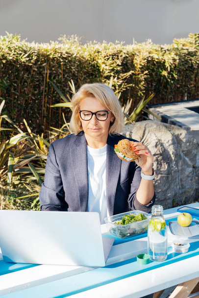 Middle-aged businesswoman having lunch at the park outdoor cafe, picnic area and working on laptop during her break. Balanced healthy diet lunch box. Unhealthy eating habits. Bad work-life balance - Fotografie, Obrázek