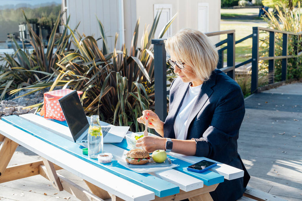 Middle-aged businesswoman having lunch at the park outdoor cafe, picnic area and working on laptop during her break. Balanced healthy diet lunch box. Unhealthy eating habits. Bad work-life balance - 写真・画像