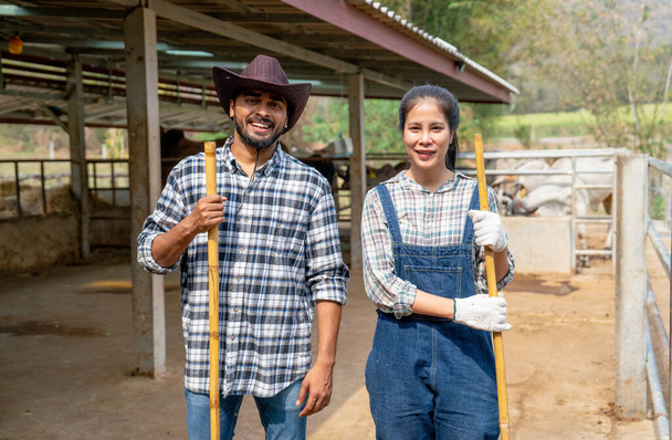 Retrato de hombre y mujer asiáticos agricultores llevan escoba y mirar a la cámara con sonrisa también de pie delante de establo de vaca mirada relajarse después de limpiar la zona. - Foto, Imagen