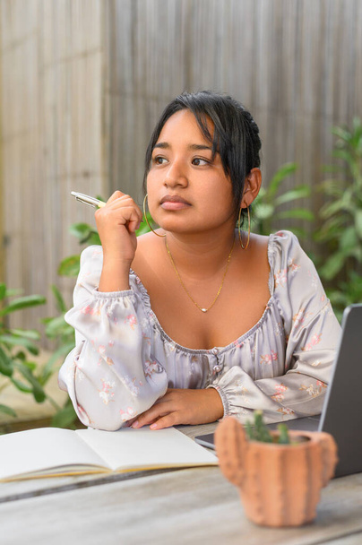 Young latin Mexican female in casual clothes sitting at table with laptop and taking notes in notebook while working on project - 写真・画像