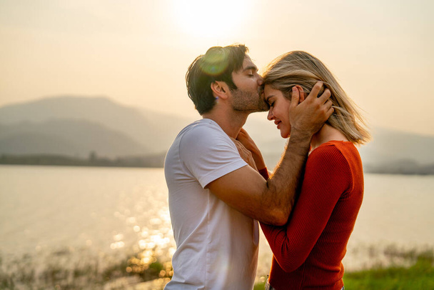 Zamknij się kaukaski mężczyzna pocałunek na czole pięknej kobiety z zachodem słońca światło w pobliżu jeziora i wyglądają romantycznie dla pary miłość pobyt razem. - Zdjęcie, obraz