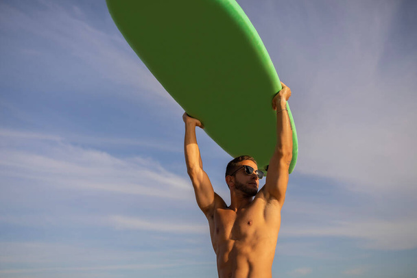 Young Muscular Male Surfer Holding Surfboard Over Head And Looking Aside, Handsome Athletic Millennial Guy In Stylish Sunglasses Posing Over Sky Background, Enjoying Summer Surfing, Copy Space - Фото, изображение