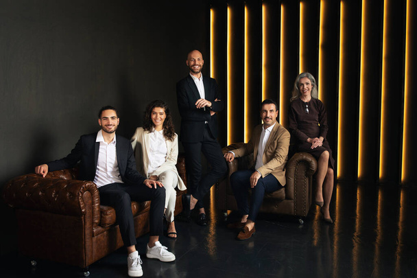 成功したビジネス会社。ブラウンソファとアームチェアに座っている5人の幸せなラテンビジネスマンとビジネスマンの女性グループ,高級ダークオフィスの屋内でポーズ.プロフェッショナルマネジメントチーム - 写真・画像