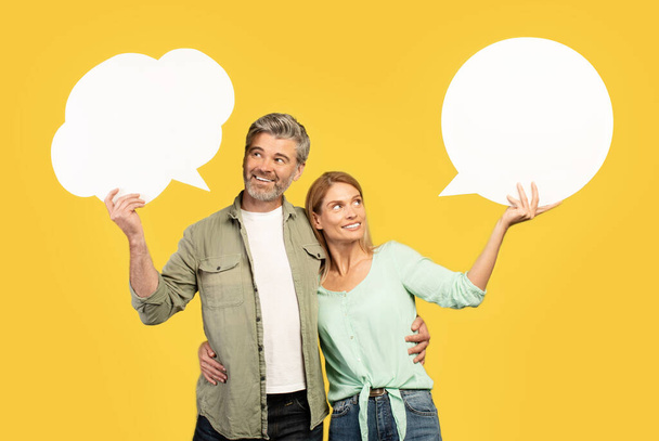 Euroopan keski-ikäinen pari, jolla on tyhjiä puhekuplia ja syleilyä, poseeraa keltaisella studiotaustalla, mallina muotoilulle. Onnelliset puolisot tyhjällä sanalla pilvet - Valokuva, kuva