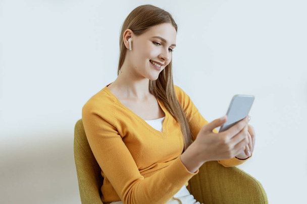 Młoda kobieta nosząca słuchawki Słuchanie audiobooka przez telefon, siedząca w wygodnym fotelu w pobliżu białej ściany w pomieszczeniach. Happy Blonde Lady relaks Korzystanie z muzyki Online - Zdjęcie, obraz