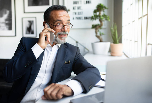 Позитивный зрелый европейский мужчина в костюме работает за ноутбуком, делая телефонный звонок в современном офисном интерьере. Мбаппе, бизнесмен, бизнес с устройствами, финансы, маркетинг и общение с клиентом - Фото, изображение