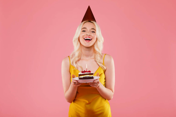Muotokuva innoissaan nainen yllään puolue hattu, juhlii syntymäpäivää ja tilalla pala kakkua kynttilän päälle vaaleanpunainen studio tausta. Onnellinen nainen nauttii hänen b-päivä - Valokuva, kuva