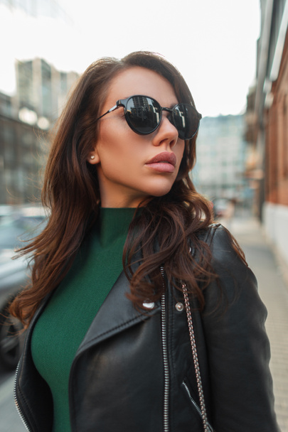 Mujer urbana hermosa joven de moda con gafas de sol vintage fresco en traje de calle rock de moda con chaqueta de cuero negro con estilo y paseos vestido verde en la ciudad - Foto, imagen