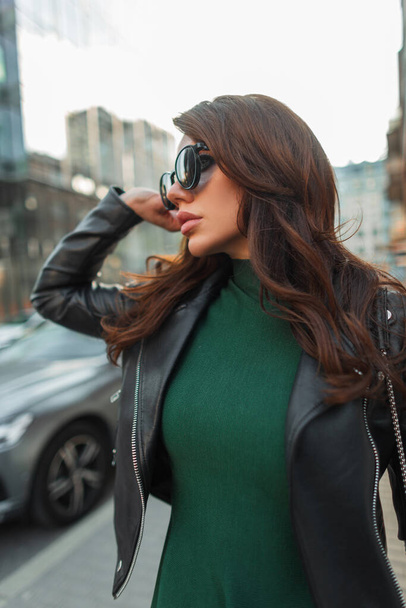Модная молодая красивая шикарная женщина в модных солнцезащитных очках в модной одежде с зеленым платьем и черной кожаной курткой гуляет по городу - Фото, изображение