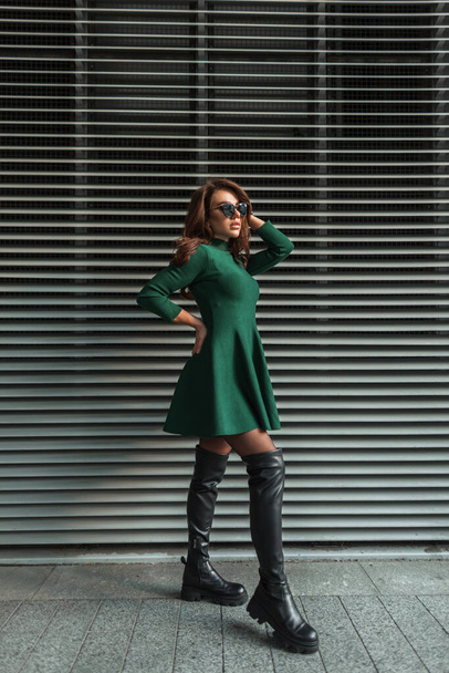 fashionable beautiful young girl with trendy sunglasses in a fashion green dress and stylish high leather shoes stands near the metal lines wall on the street - Photo, image