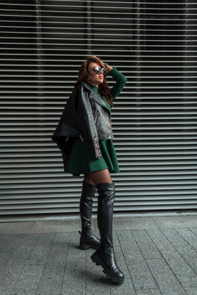 Cool hermosa mujer sexy con estilo con gafas de sol en moda elegante traje de rock con vestido verde y chaqueta de cuero negro con zapatos altos posando cerca de la pared de metal al aire libre - Foto, imagen