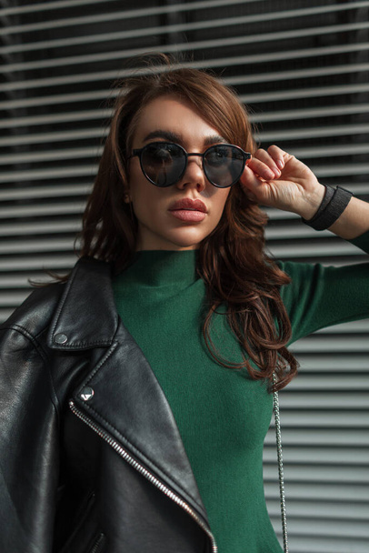 Pretty cool stylish beauty woman in trendy fashion clothes with black leather rock jacket puts on sunglasses and walks on the street near the metal lines background - Foto, imagen