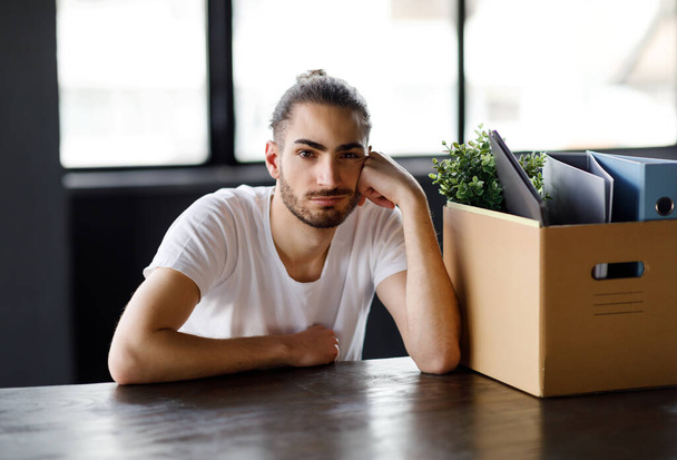 Licenciement. licencié arabe employé homme assis à la table avec son appartenance dans la boîte en carton sur le lieu de travail, regardant la caméra posant dans l'intérieur de bureau moderne. Concept du problème du chômage - Photo, image