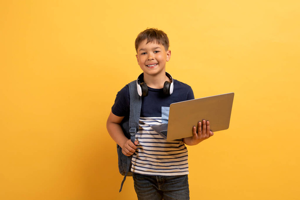 Wesoły przystojny chłopak ze szkoły kaukaskiej w luźnym stroju z plecakiem i bezprzewodowymi słuchawkami trzymającymi i używającymi laptopa stojącego na żółtym tle. Edukacja i technologie - Zdjęcie, obraz