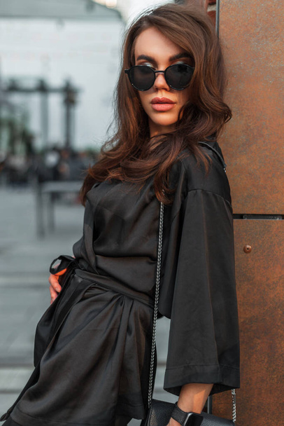 Moda gözlüklü, şık, siyah takım elbiseli, şık, genç ve zarif bir kadın model. Şehirde bir duvarın yanında duruyor. - Fotoğraf, Görsel