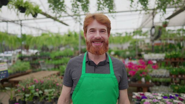 Portrait of a happy redhead male worker wearing Green apron standing inside flower shop. Local business concept of man employee - Fotó, kép