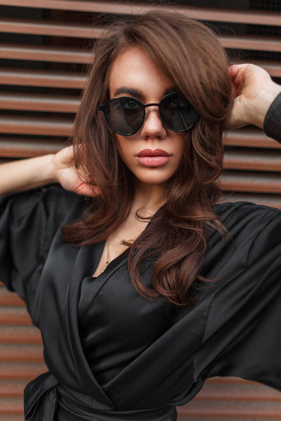 Retrato femenino al aire libre de una hermosa mujer morena elegante con gafas de sol de moda cool en un vestido negro cerca de una pared de metal en la ciudad - Foto, Imagen
