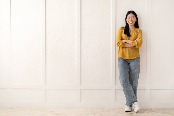 Positivo atraente jovem coreana vestindo roupa elegante casual posando sobre a parede vazia em branco em casa, olhando e sorrindo para a câmera. Asiático senhora procurando novo apartamento, realocando - Foto, Imagem
