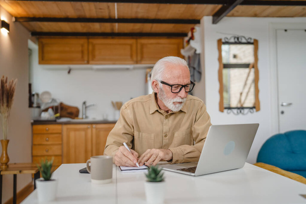 Egy idős, fehér férfi nagyapa szakállal és szemüveggel otthon számítógépen dolgozik. - Fotó, kép