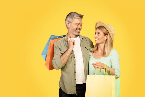 Excited european middle aged couple posing together with colorful shopper bags showing their purchases, enjoying shopping during huge sales on yellow studio background - Foto, Bild
