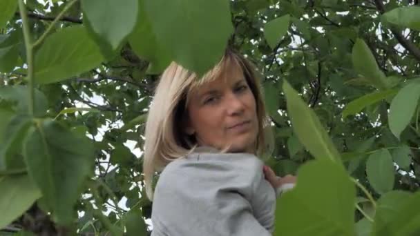 Bella donna tra le foglie dell'albero posa per la fotocamera. Una donna con i capelli chiari - Filmati, video