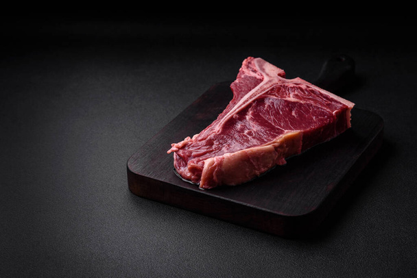 Saftiges rohes Rindfleisch-T-Bone-Steak mit Salz, Gewürzen und Kräutern auf texturiertem Betonhintergrund - Foto, Bild