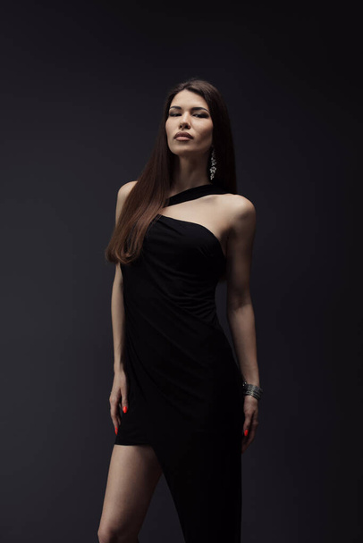Sensual young female in stylish black dress standing in studio and looking at camera on black background during photo shoot against dark backdrop - Photo, image