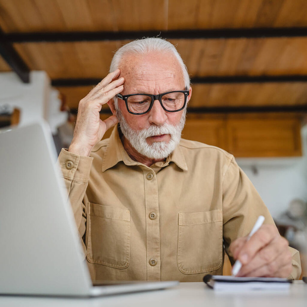 Een senior Kaukasische man grootvader met baard en bril werk op laptop gebruik computer thuis oplossen probleem bezorgd frustratie - Foto, afbeelding