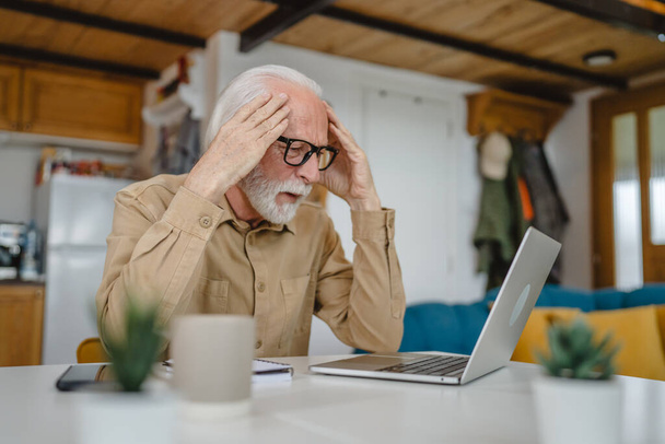 Un hombre mayor caucásico abuelo con barba y anteojos de trabajo en el ordenador portátil de uso en casa resolver el problema preocupado frustración - Foto, Imagen