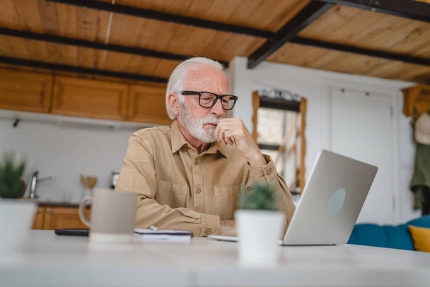 Один старший кавказький чоловік дід з бородою та окулярами працює над ноутбуком, використовуючи комп'ютер вдома, вирішуючи проблему, стурбований розчаруванням
 - Фото, зображення
