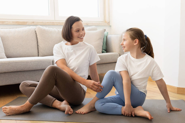 Sports at home together. Smiling millennial caucasian lady in sportswear and teenage daughter practice yoga, training in living room interior. Body care and fitness, support mom - Foto, imagen