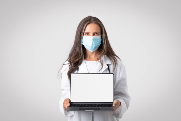 Professional senior doctor woman in white coat, wearing protective mask showing laptop with blank white screen, posing on gray background, mockup. Treatment, remote service, advice, ad, offer - Photo, Image