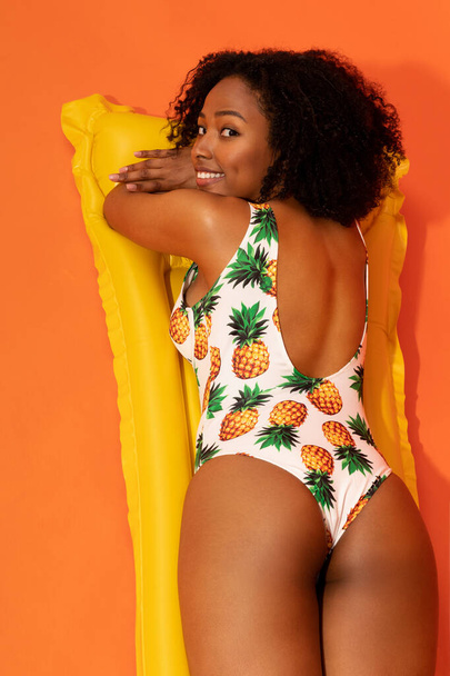 Nyugodt boldog mosolygós vonzó fiatal afro-amerikai nő visel egyrészes fürdőruha feküdt sárga úszó felett narancs háttér, van napozás, szolárium, függőleges stúdió lövés - Fotó, kép