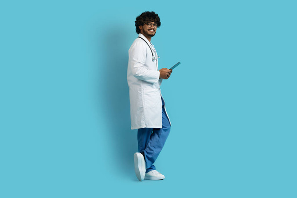 Nykyaikainen teknologia terveydenhuollossa. Onnellinen komea nuori intialainen kaveri lääketieteen työvaatteet lääkäri käyttää digitaalinen tabletti eristetty sinisellä pohjalla, doc chattailuun potilaan kanssa, kopioi tilaa - Valokuva, kuva