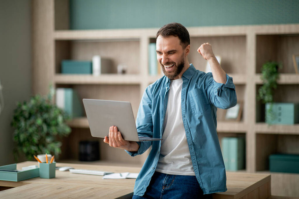 Business Success. Young Happy Businessman Celebrating Success With Laptop In Office, Overjoyed Male Entrepreneur Shaking Fist And Shouting Yes, Standing With Computer Near Desk Indoors, Copy Space - Fotoğraf, Görsel