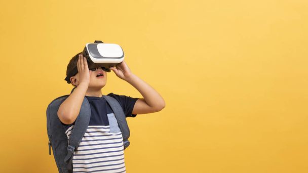 Schooler boy in casual outfit with backpack behind his back using VR glasses isolated on yellow background, virtual reality experience, copy space, banner. Modern technologies in kids education - Photo, Image