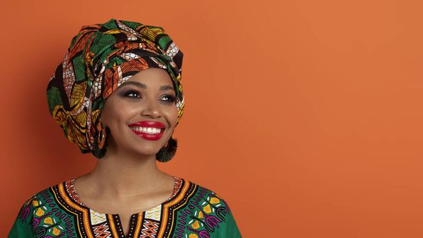 コピースペースと笑顔、パノラマを見て明るいメイクを身に着けている伝統的なアフリカの衣装で幸せな陽気な美しい若い黒人女性のクローズアップスタジオショット。いい提案だ - 写真・画像